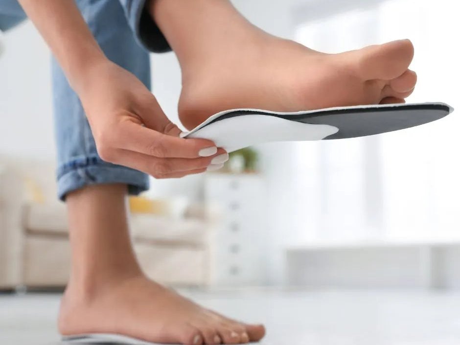 Two hands holding a Redi-Thotics orthotic up against a bare foot in a bright room, checking how the arch lines up.